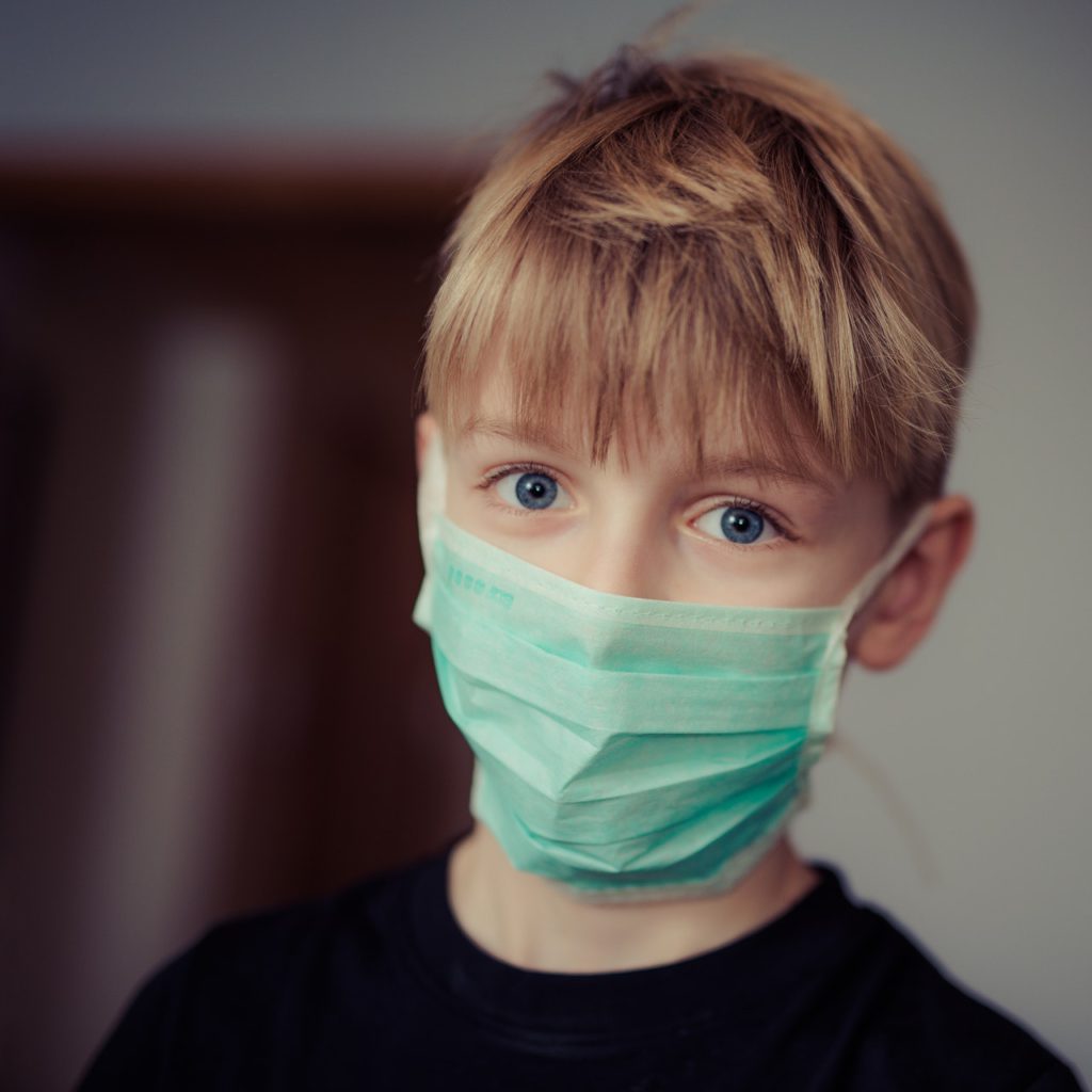 blonde haired white boy wearing a green tinted surgical mask facing camera