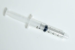 Photo looking down on a fat clear plastic syringe with a blunt end (no needle) with the plunger pulled partially back against a neutral light surface.