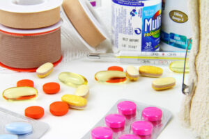 Stock photo including a roll of ACE bandage tape and a glass mercury thermometer in the back ground, various colorful capsules and tablets and pills strewn in the foreground, and an over the counter pill bottle in the background representing a variety of standard remedies in western allopathic medicine.
